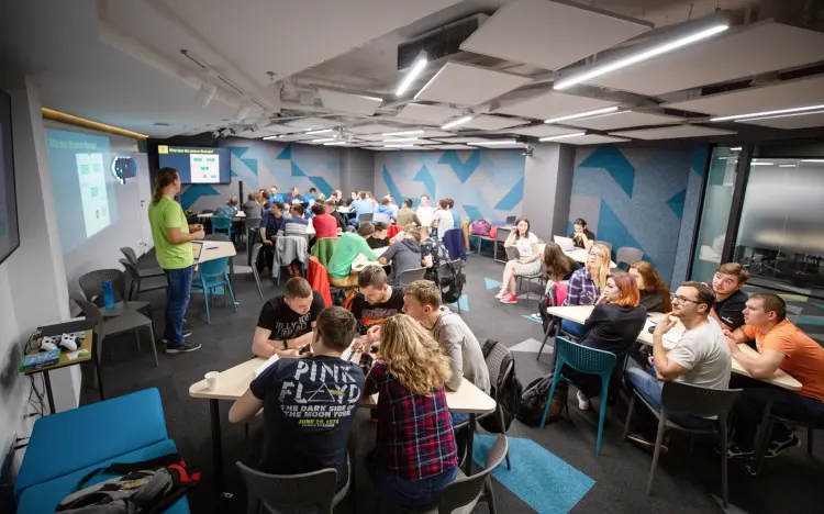 Classroom setting with groups at tables, a presenter stands at the front near a projected screen